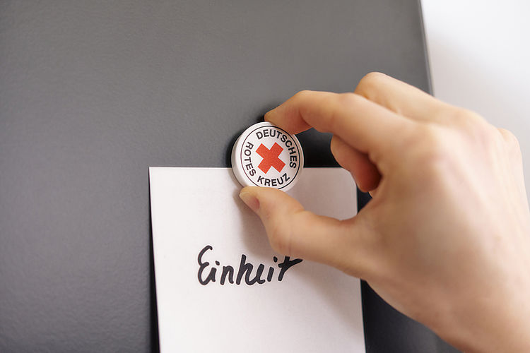 Symbolfoto Magnet mit DRK - Logo befestigt Zettel mit dem Schriftzug Grundsatz ‚Einheit‘ auf einer Tafel