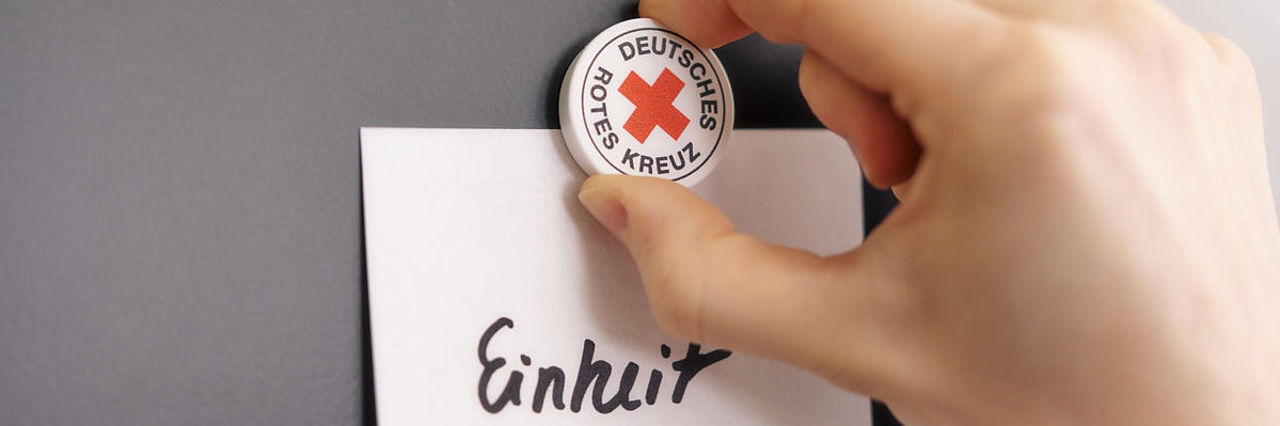 Symbolfoto Magnet mit DRK - Logo befestigt Zettel mit dem Schriftzug Grundsatz ‚Einheit‘ auf einer Tafel