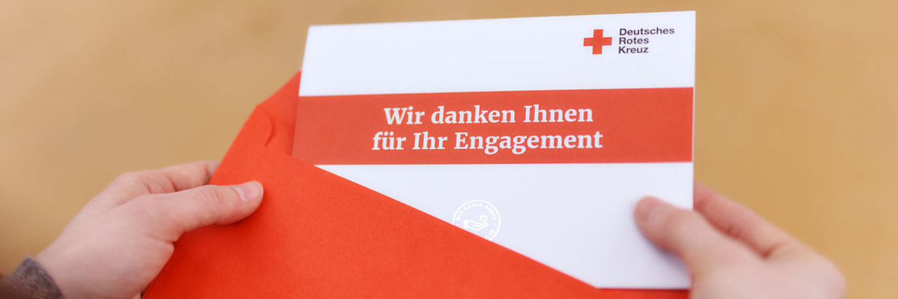 Symbolfoto Empfängerin nimmt eine Grußkarte des DRK mit der Aufschrift "Wir danken Ihnen für Ihr Engagement" aus dem Briefumschlag