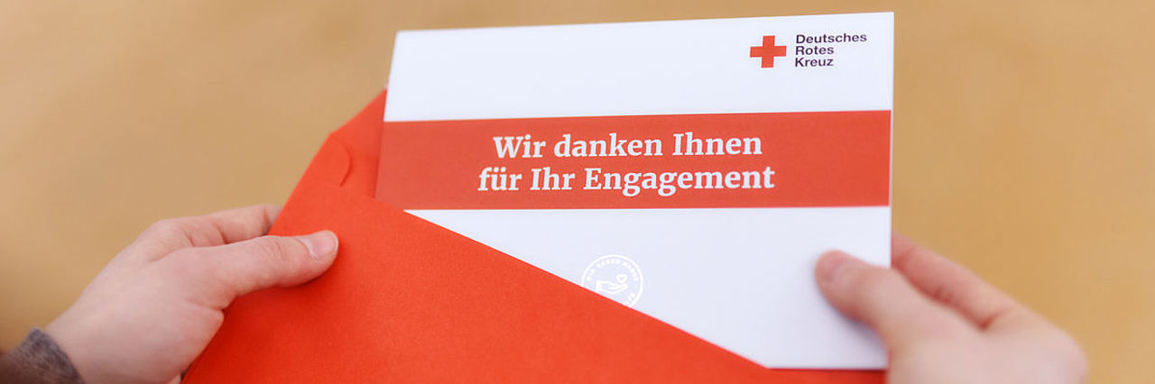 Symbolfoto Empfängerin nimmt eine Grußkarte des DRK mit der Aufschrift "Wir danken Ihnen für Ihr Engagement" aus dem Briefumschlag